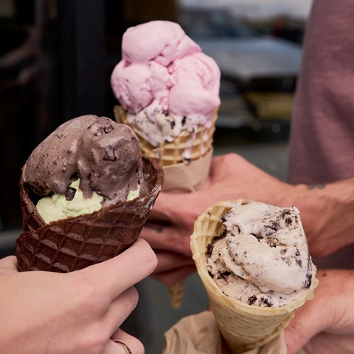 Photograph of three ice creams being held by three hands. The ice creams are pink, white, and brown.