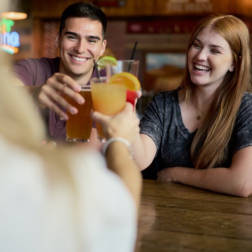Photograph people sitting around a table holding their drinks up mid-cheers. They are in a bar with wood walls. 