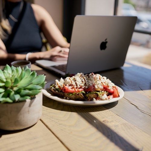 Photograph of some avocado toast on a table, in front of a person working on a laptop. There is a succulent on the table. 